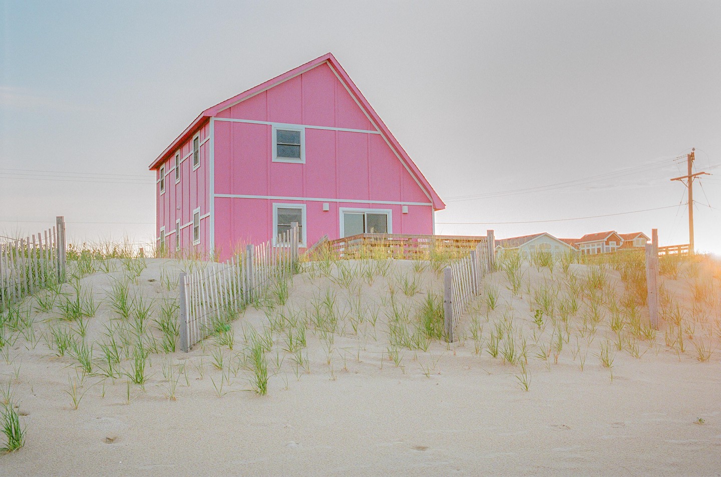 A bright pink house stands behind a sand dune with a wooden fence and sparse green grass.
