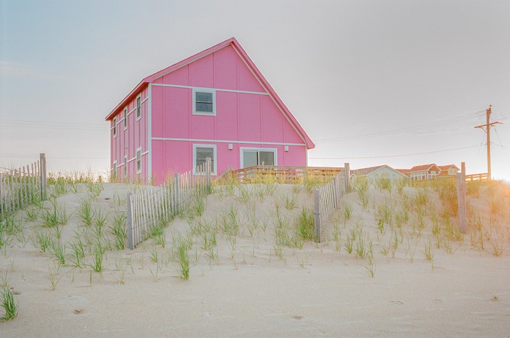 A bright pink house stands behind a sand dune with a wooden fence and sparse green grass.