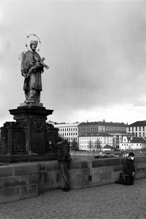 Two men are near a statue of a saint holding a crucifix on a bridge with buildings in the background.