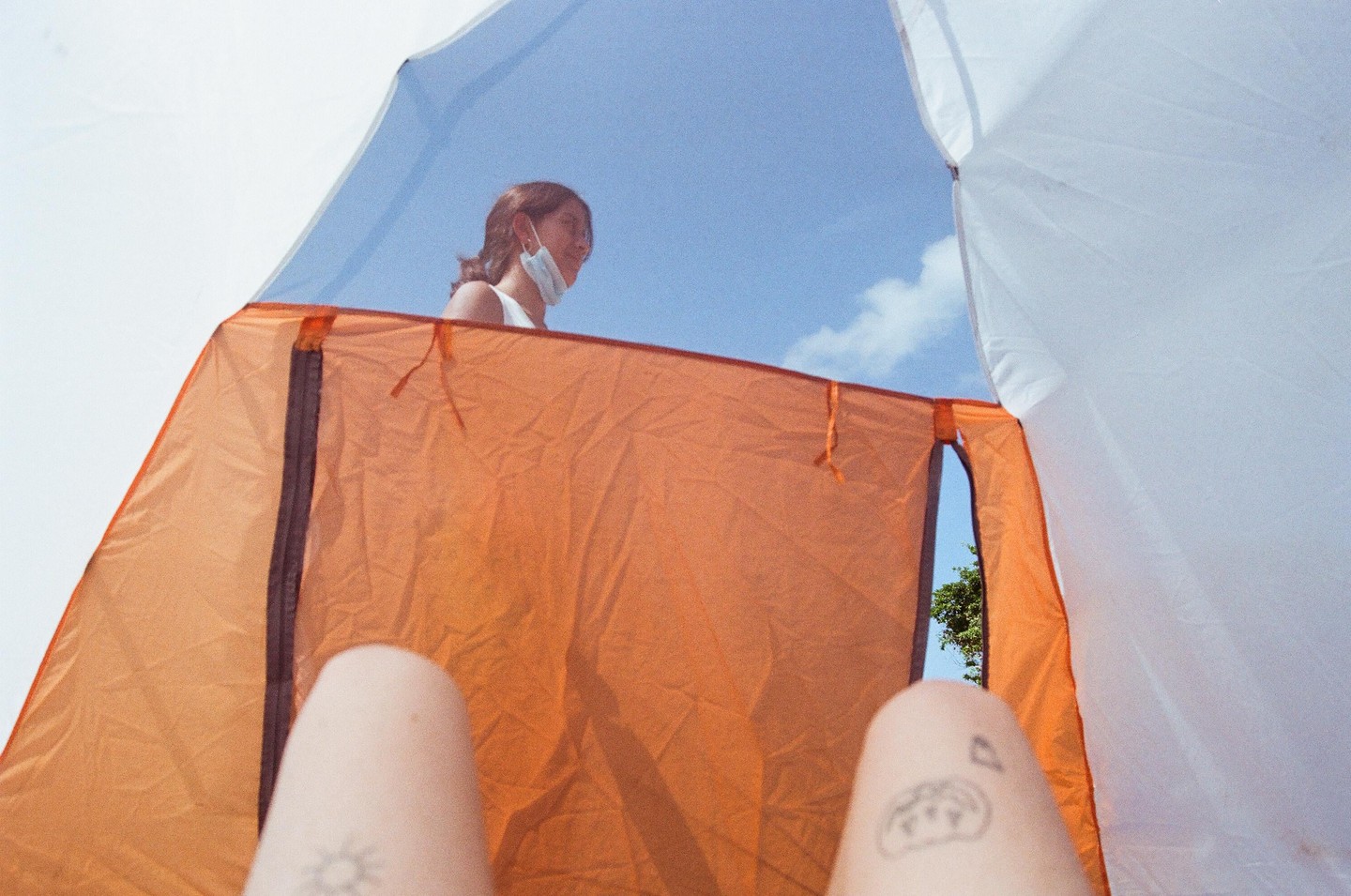 A woman with a face mask stands outside a tent, viewed from inside the tent with two tattooed knees in the foreground.