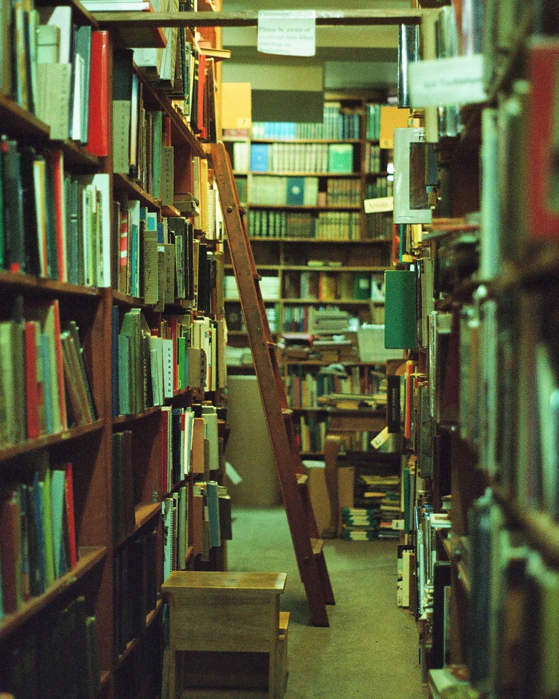 A narrow aisle in a library is lined with bookshelves and features a wooden stool and a tall ladder.