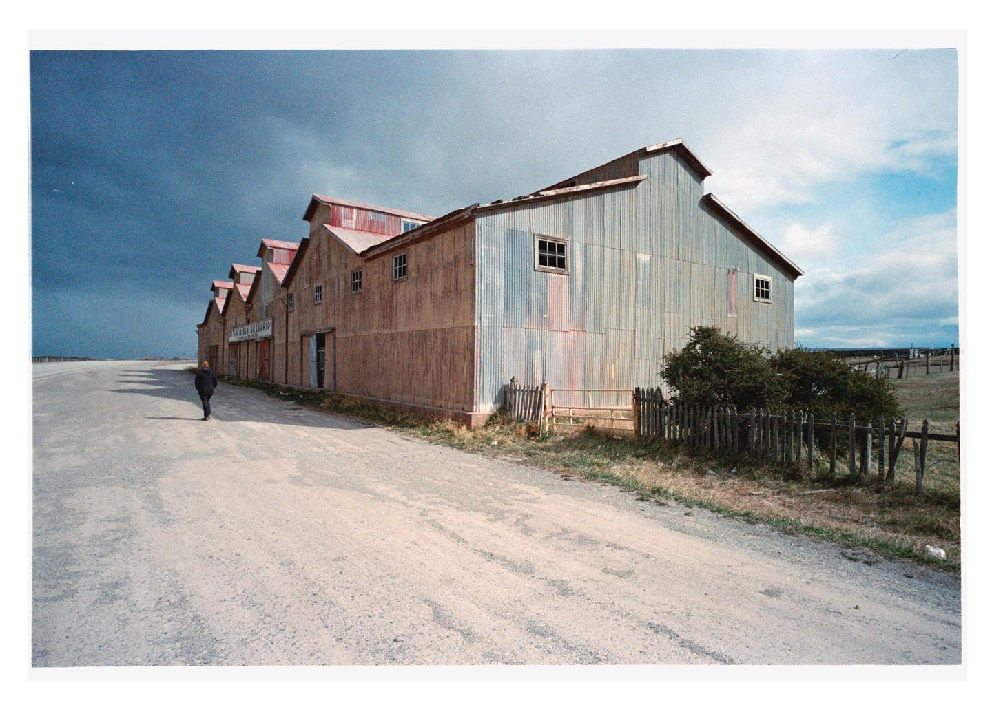 A man walks along a dirt road next to a long, corrugated metal building under a dramatic sky.