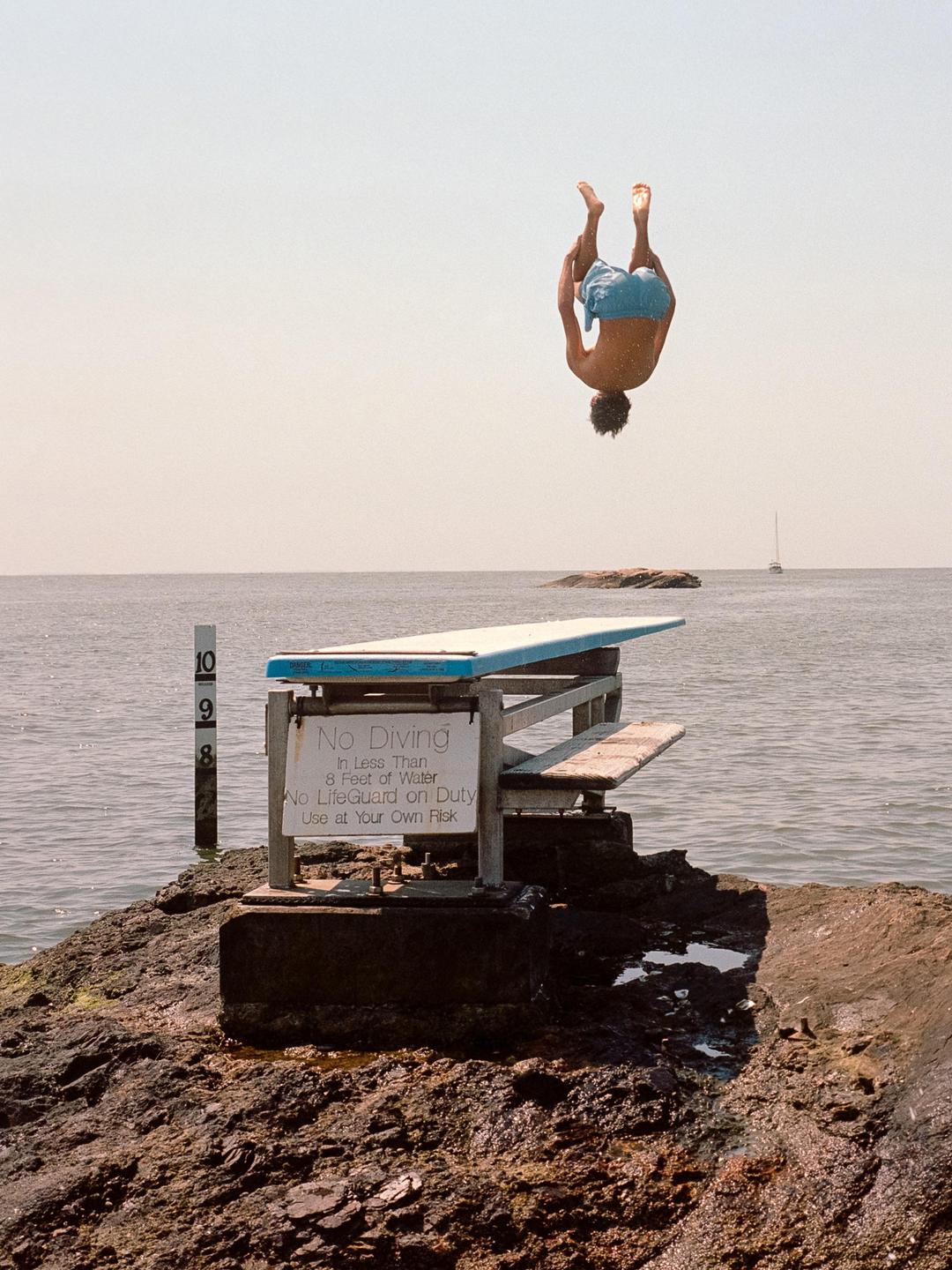 A man performs a backflip off a diving board over the ocean, with a "No Diving" sign visible on the board's support structure.