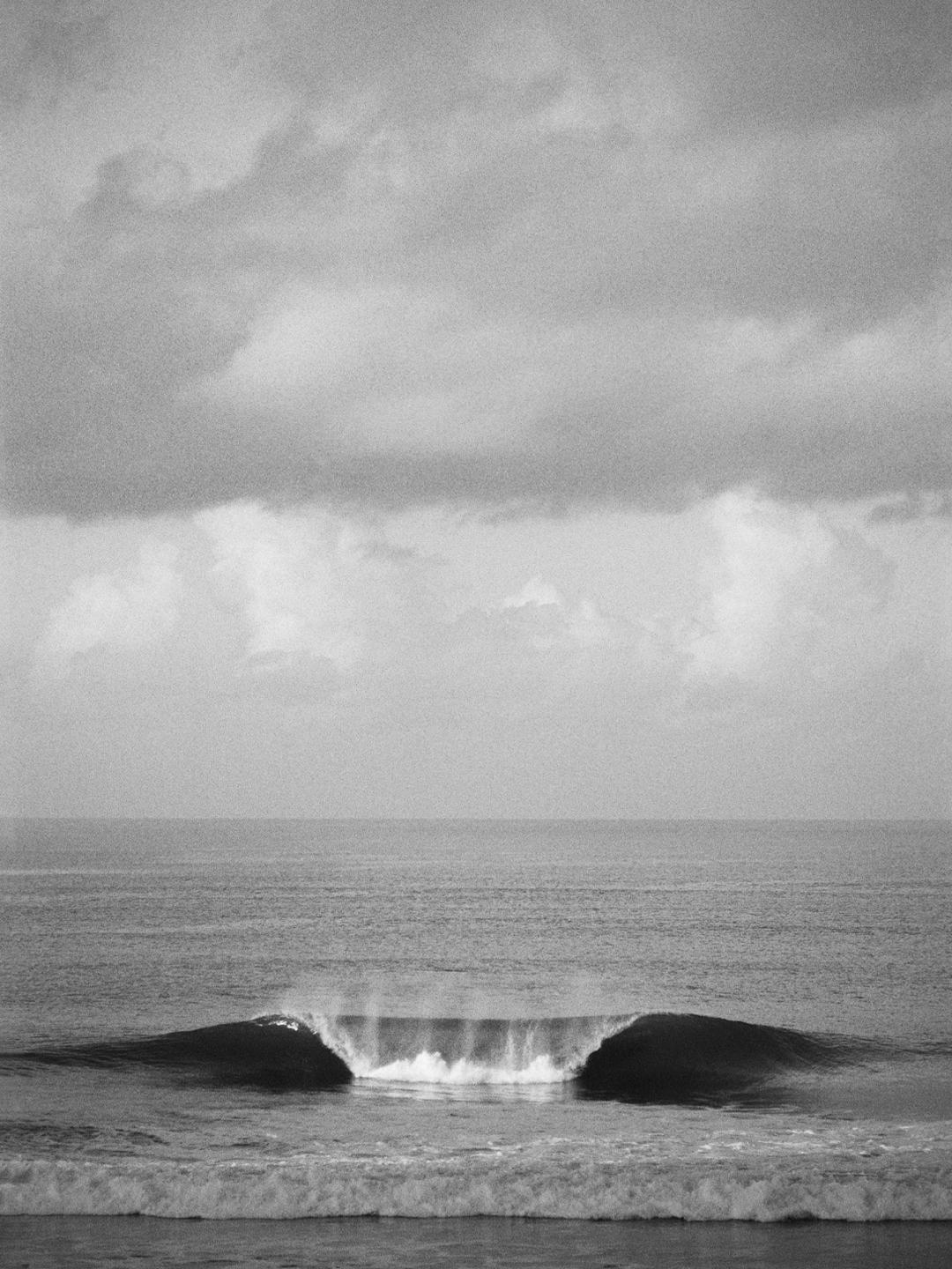 A large wave breaks in the ocean under a cloudy sky.