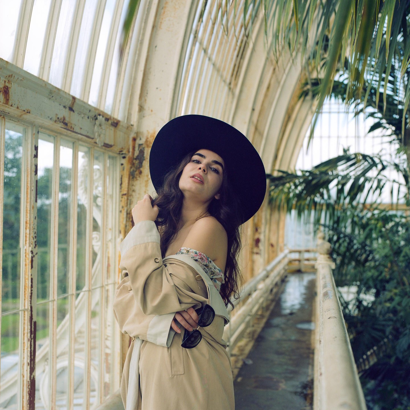 A woman in a wide-brimmed hat and trench coat poses in a greenhouse with lush foliage.