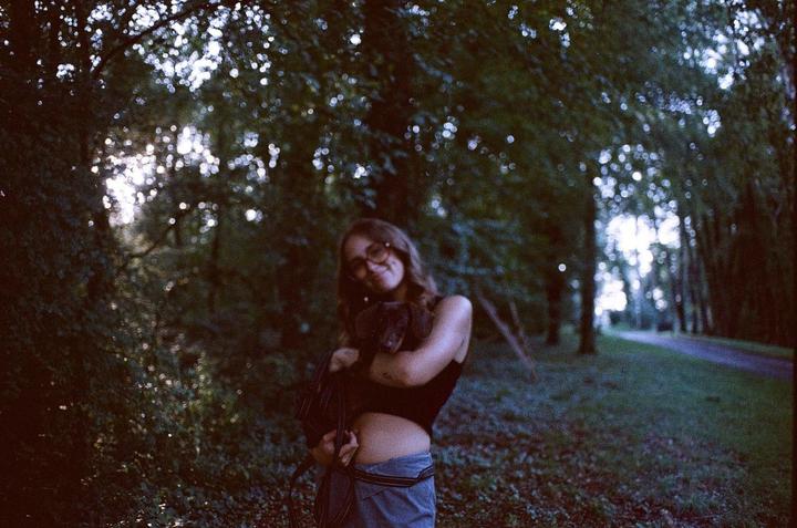 A woman holds a dachshund in a forest with bright light filtering through the trees.