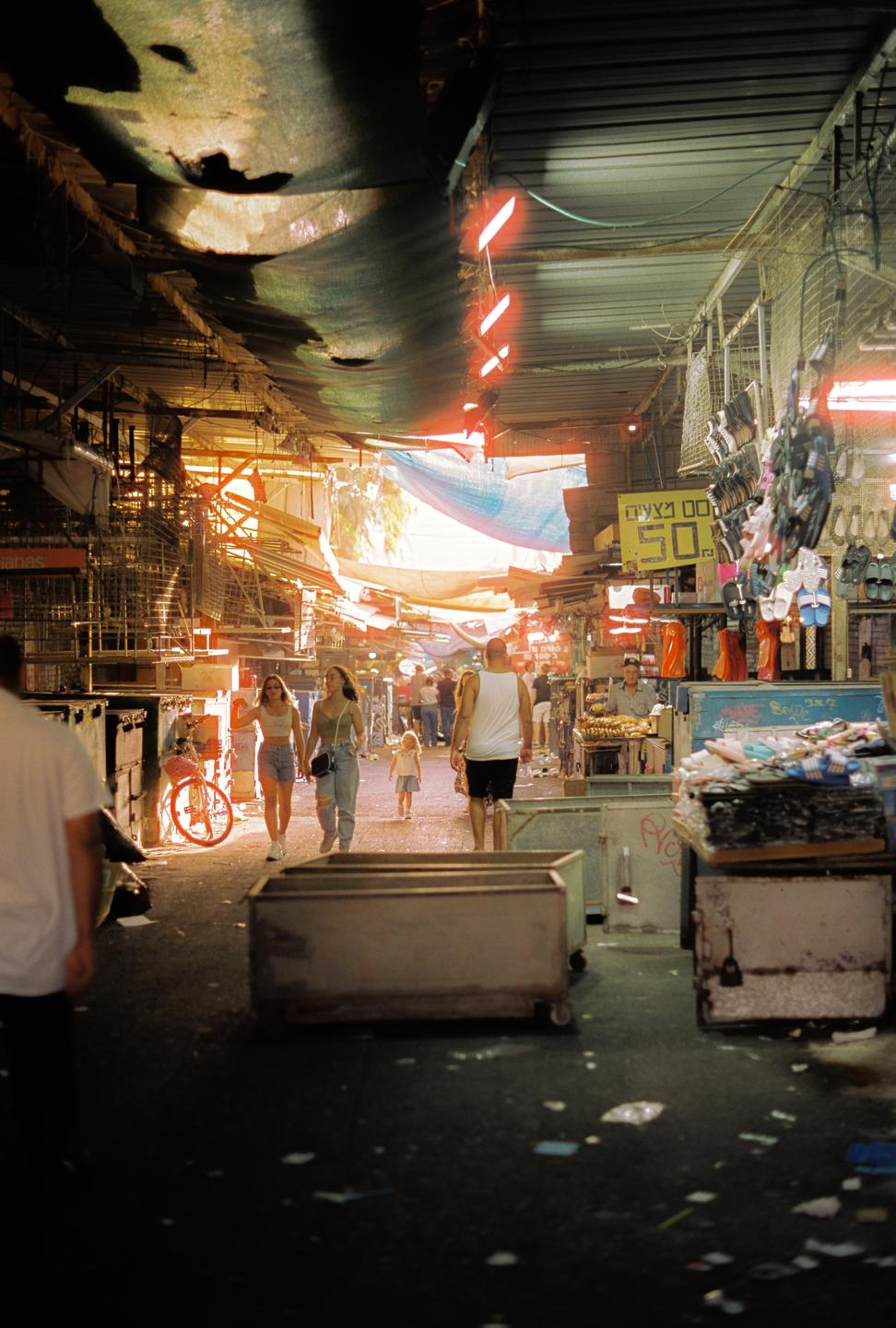 People walk through a bustling market with stalls on either side and light streaming in from the end of the alley.