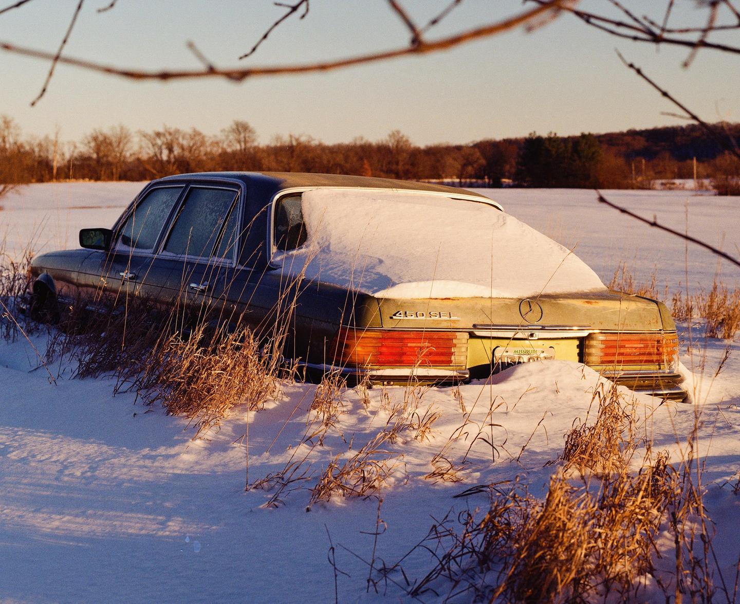 A snow-covered car is parked in a field with dry grass and trees in the background.