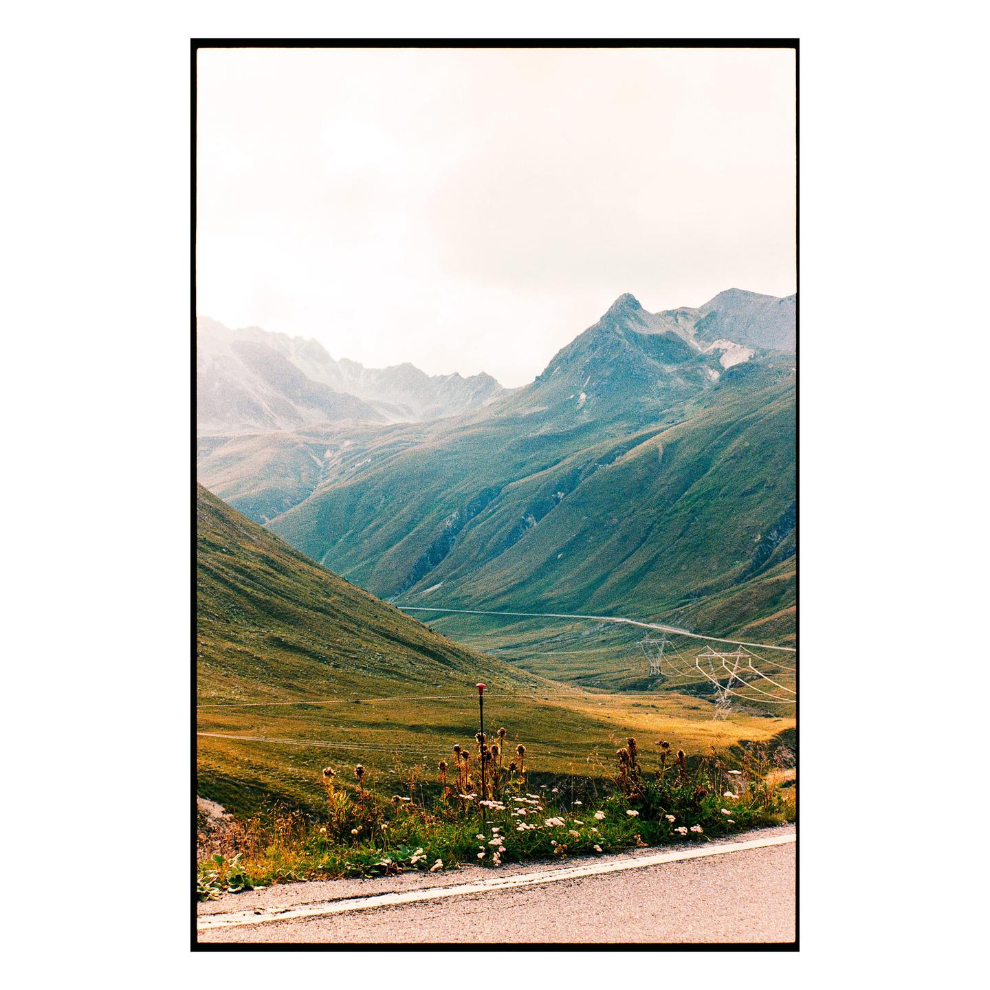A winding road cuts through a vast mountain landscape with green and golden slopes under a hazy sky.