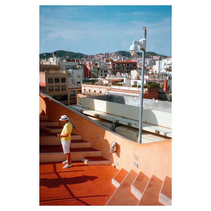 A man in a hat stands on a rooftop terrace overlooking a city.