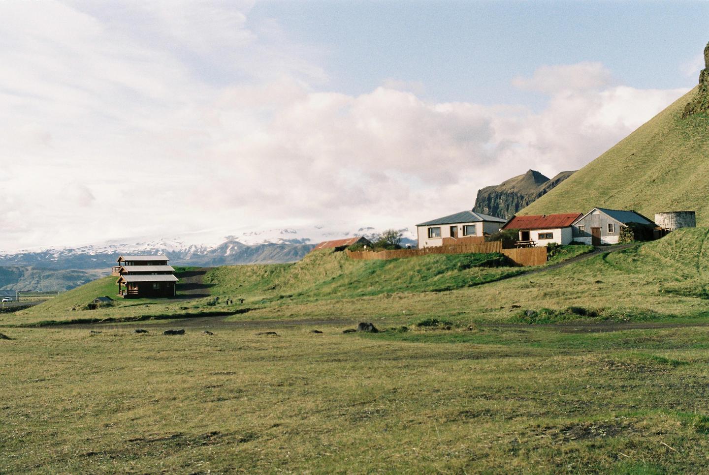 A cluster of houses sits on a green hillside with snow-capped mountains in the distance under a cloudy sky.