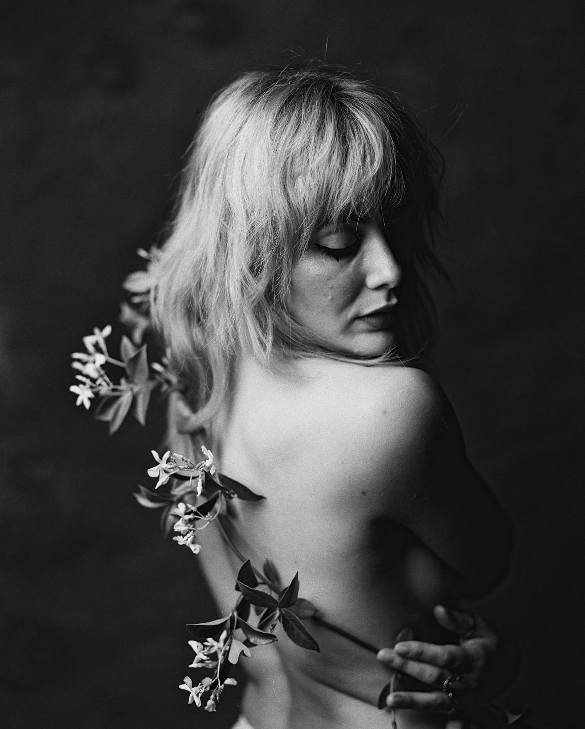 A woman with blonde hair and bare shoulders looks down, with a flowering branch draped over her back.