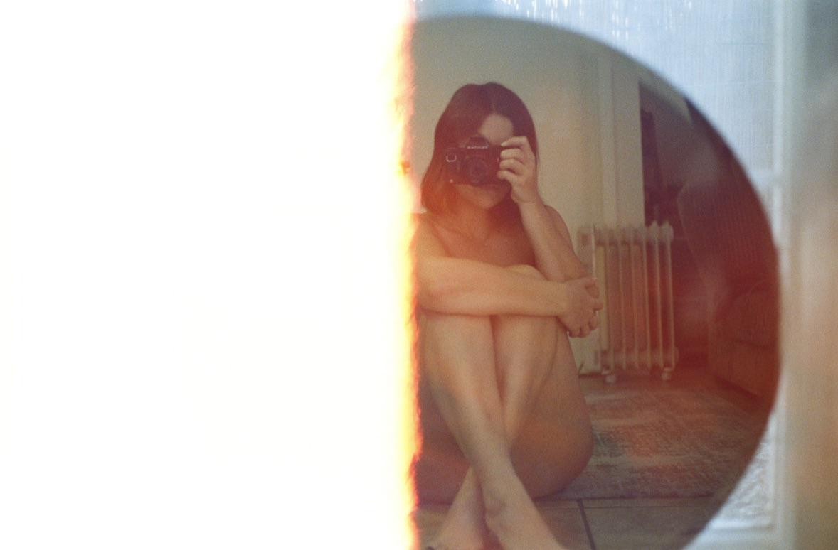 A woman sits on the floor, holding a camera to her face, reflected in a mirror with a bright light leak on the left.
