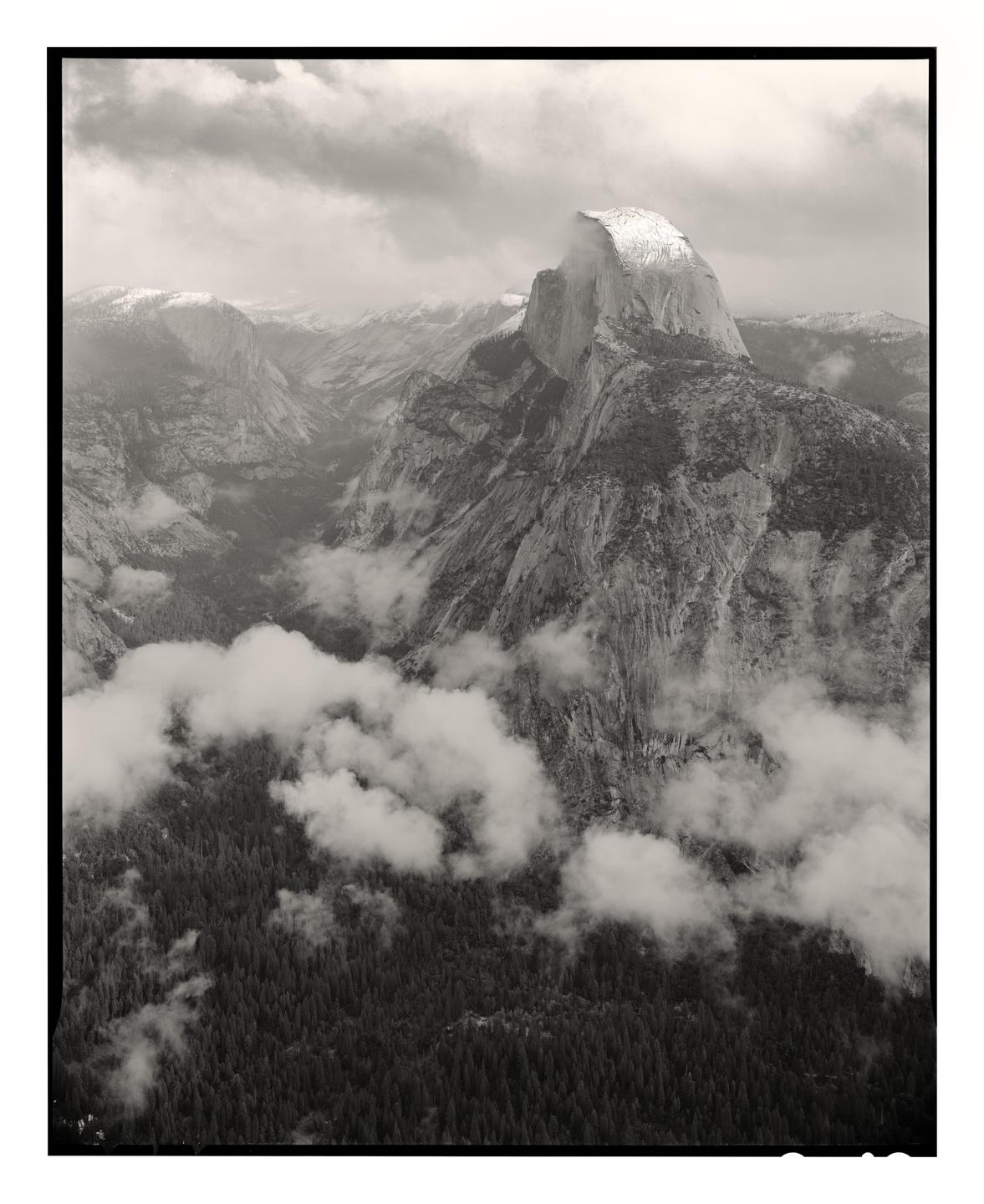 Clouds hang over a valley with a large mountain in the background and a forest in the foreground.