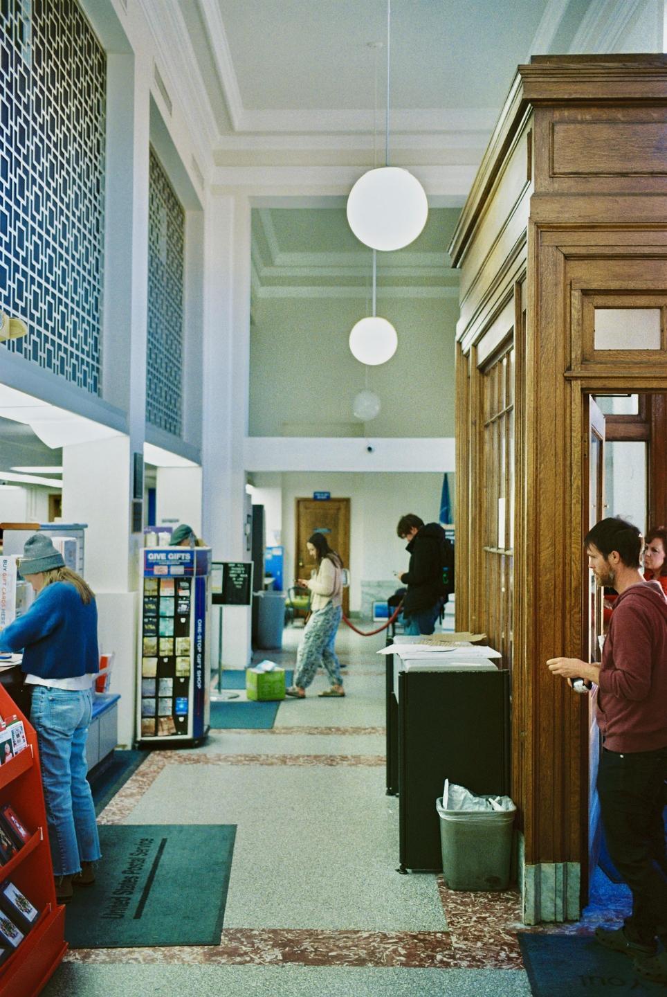 People are standing in a post office lobby with high ceilings and large windows.