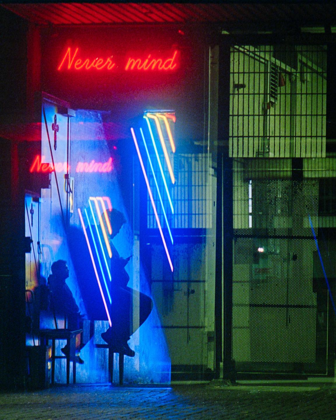 Two people sit in a doorway illuminated by red and blue neon signs that read "Never mind."