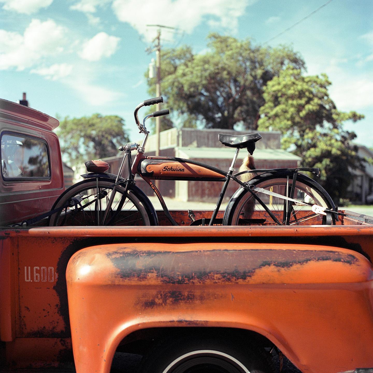 A bicycle is in the bed of an orange pickup truck under a blue sky with clouds.