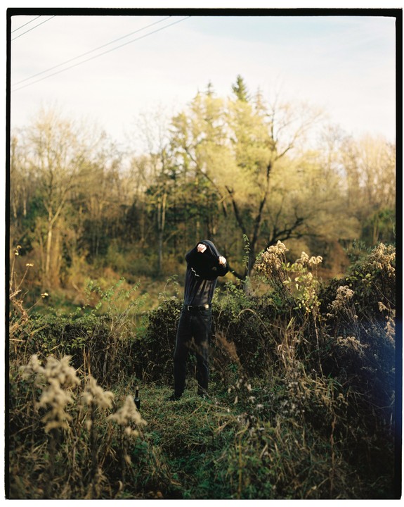 A man in a dark hoodie pulls the hood over his head in a field with trees in the background.