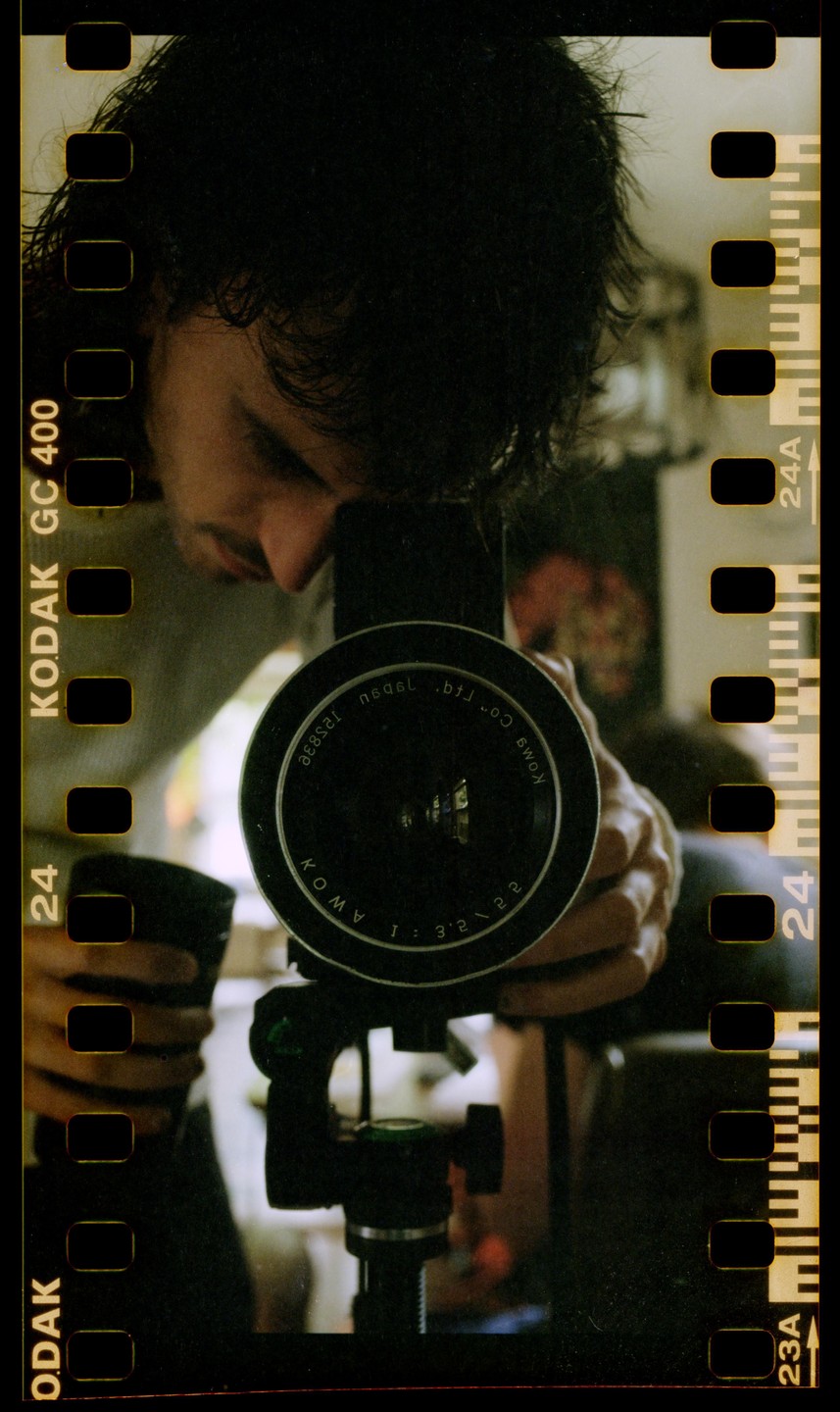 A man is reflected in a camera lens, holding the camera on a tripod.