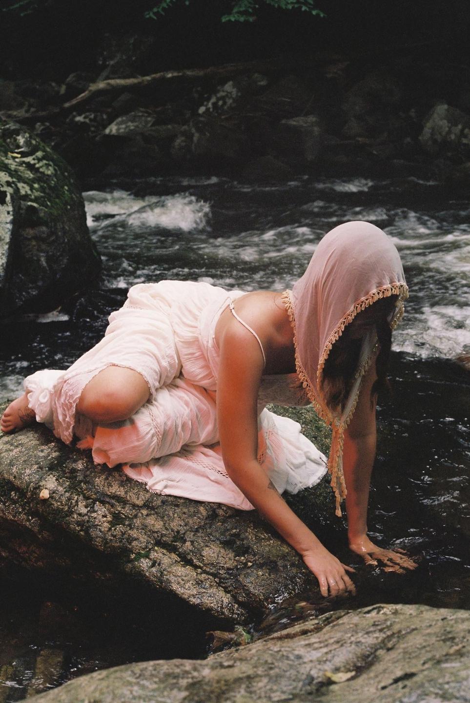 A woman in a white dress and pink head covering kneels on a rock by a river, touching the water.