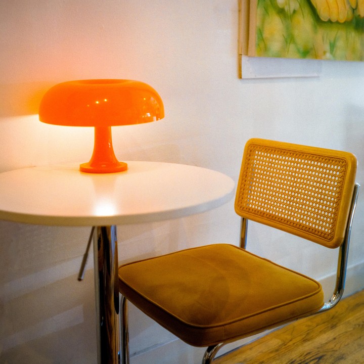 An orange mushroom lamp sits on a white table next to a chair with a cane back and mustard yellow cushion.