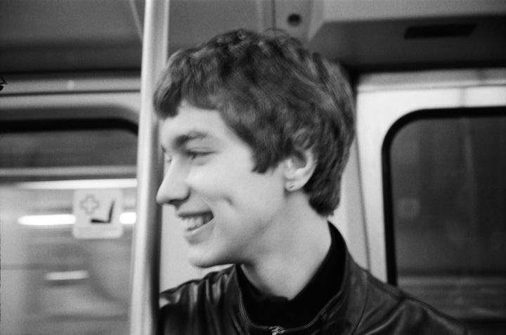 A young man with short hair and a leather jacket smiles while looking to the left.