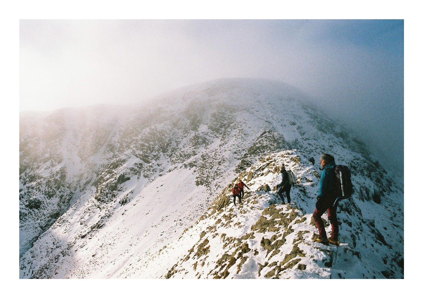 Three people hike along a snowy mountain ridge under a hazy sky.
