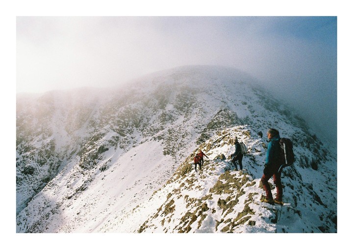 Three people hike along a snowy mountain ridge under a hazy sky.