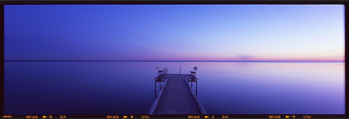 A wooden pier with two benches extends into a calm body of water under a blue and purple sky.