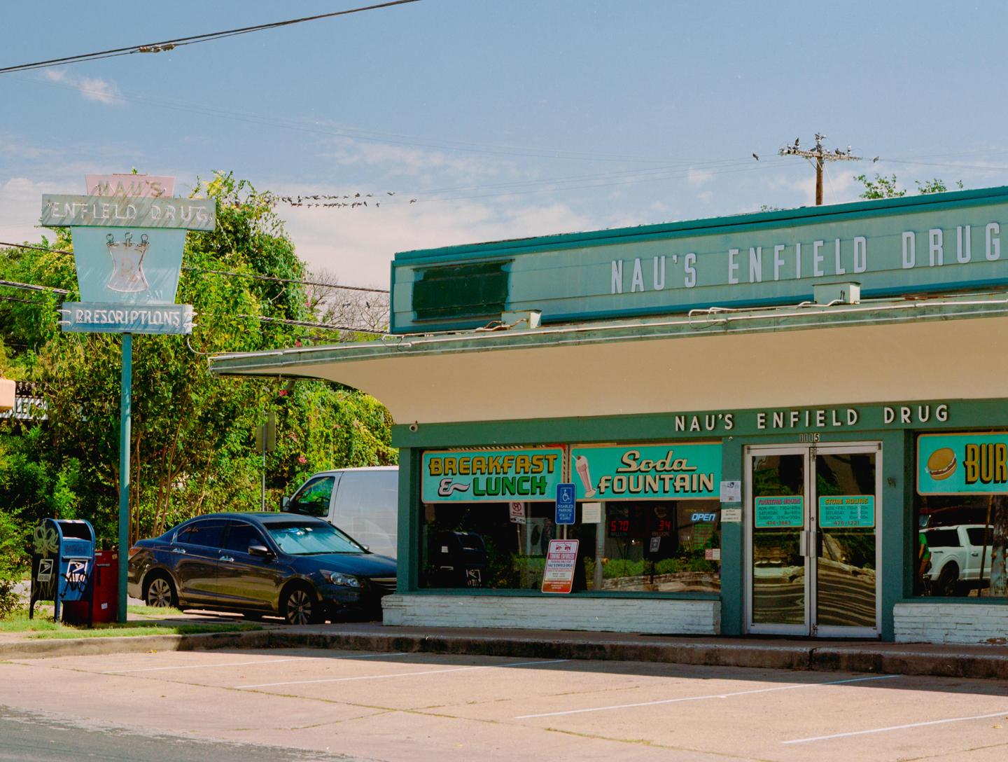 A black car is parked in front of a building with a sign that reads "Nau's Enfield Drug" and "Soda Fountain."