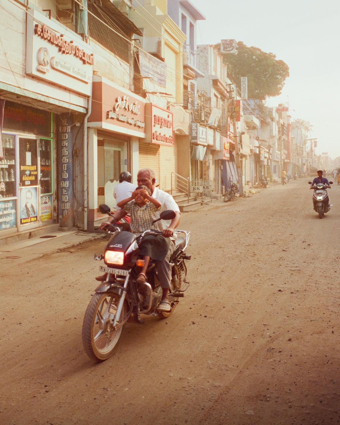 A man and a boy ride a motorcycle down a dusty street lined with buildings.