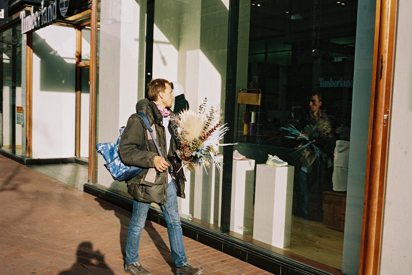 A man carrying a large bouquet of dried flowers and a backpack walks past a storefront with a reflection of himself in the window.
