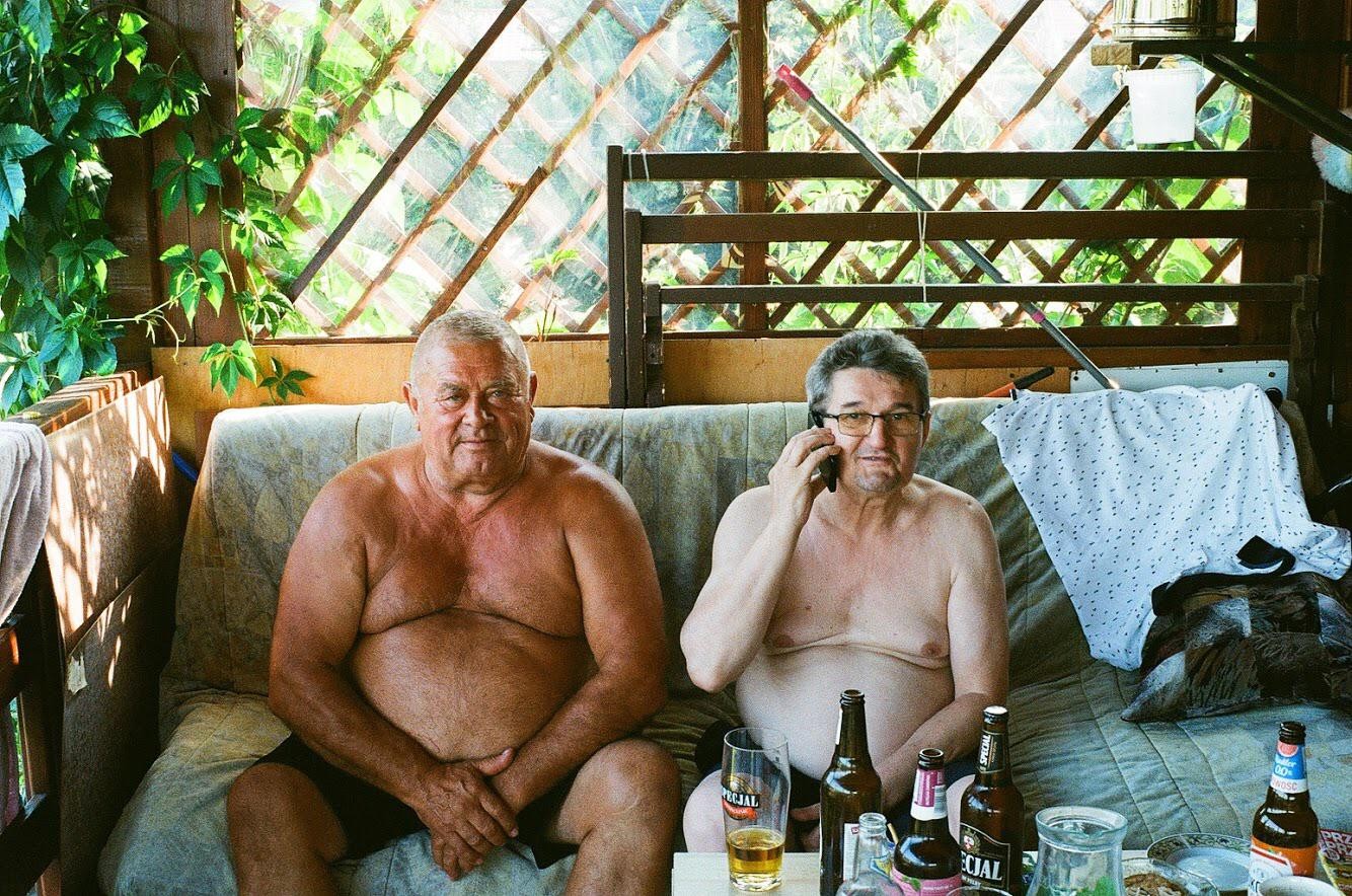 Two shirtless men sit on a couch on a porch, one talking on a cell phone, with bottles and glasses on a table in front of them.
