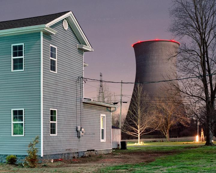 A house stands in a field with a large cooling tower in the background at dusk.