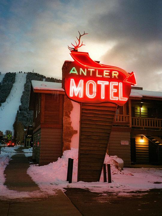 A neon sign for the Antler Motel glows red and white against a snowy mountain landscape.