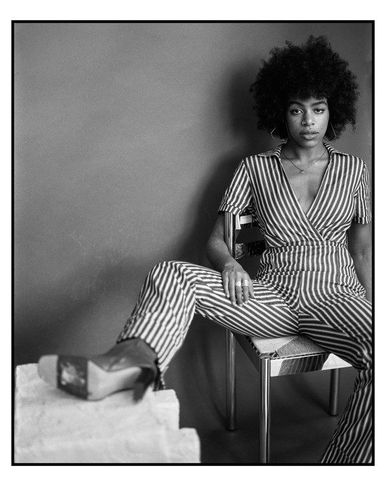 A woman with curly hair and a striped jumpsuit sits on a chair with one leg extended onto a white block.