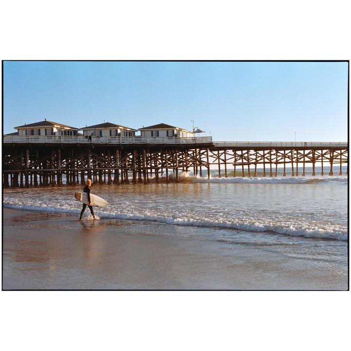 A surfer walks along the shore with a surfboard in front of a pier with buildings on it.