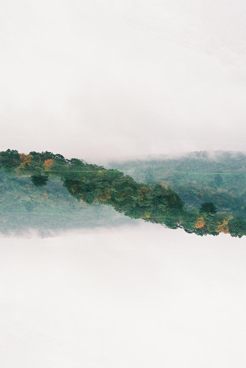A double exposure shows a forest with autumn foliage reflected in a body of water, with fog in the background.