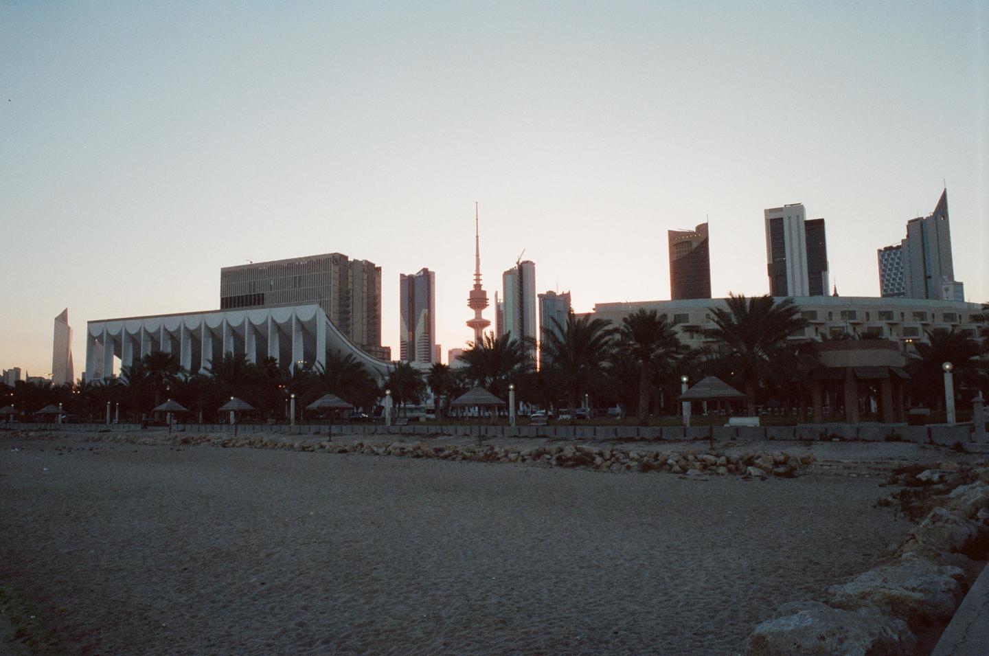 A sandy beach with rocks in the foreground looks towards a city skyline with palm trees and a prominent tower at dusk.