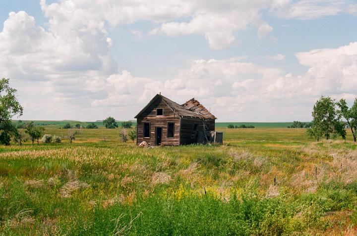 An abandoned wooden house stands in a vast field under a cloudy sky.