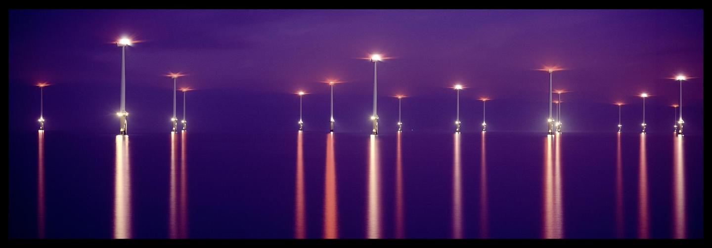 A series of offshore wind turbines are illuminated at night, casting long reflections on the water.