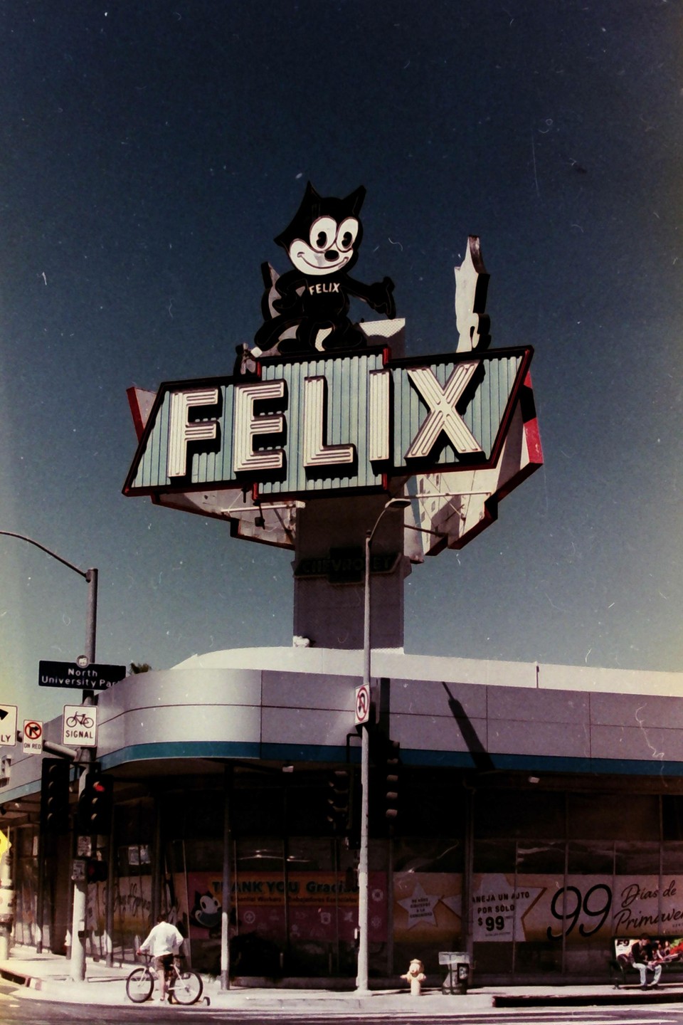 A large Felix the Cat sign sits atop a building on a street corner with a person on a bicycle passing by.