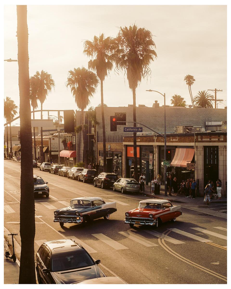 Two vintage cars drive on a street lined with palm trees and buildings under a hazy sky.