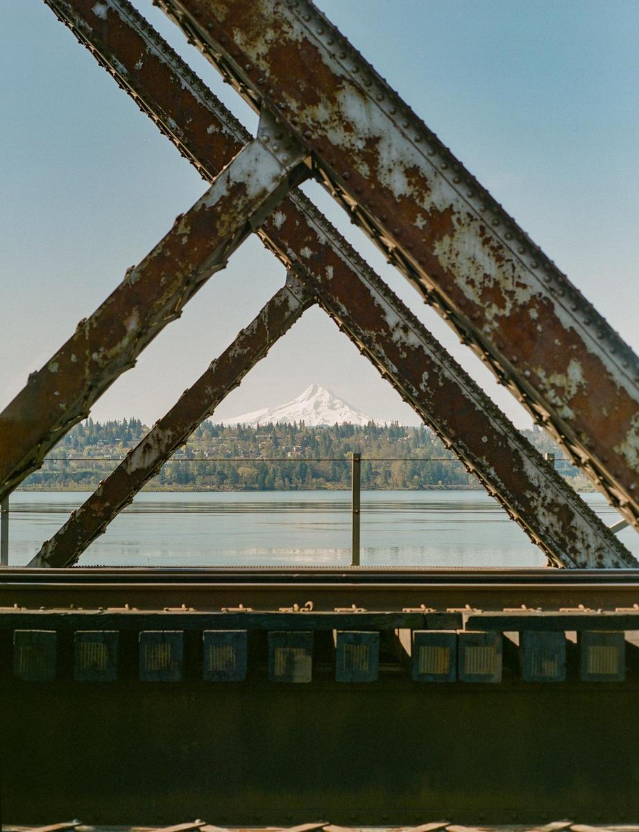 A snow-capped mountain is visible through the rusty metal structure of a bridge over water.