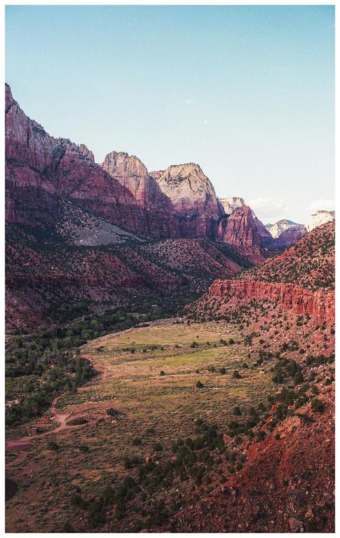 A wide valley with green trees and bushes is surrounded by red rock mountains under a clear sky.
