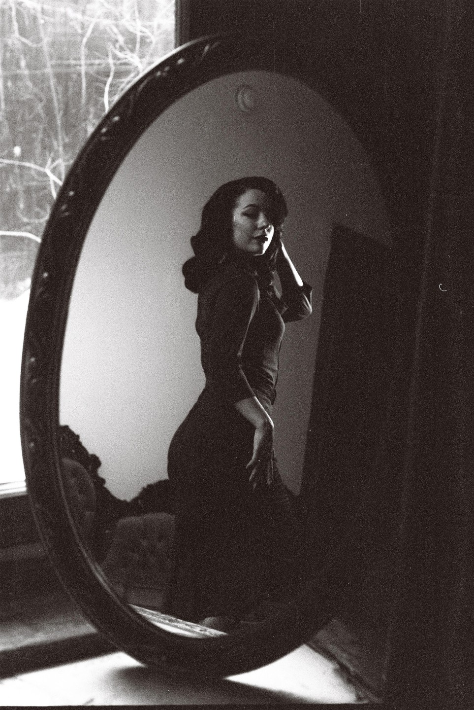 A woman in a dark dress poses in front of an oval mirror.