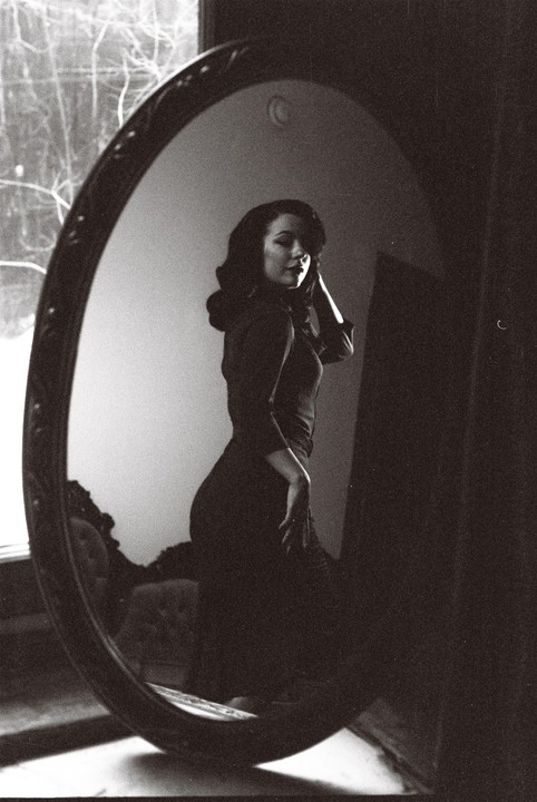 A woman in a dark dress poses in front of an oval mirror.