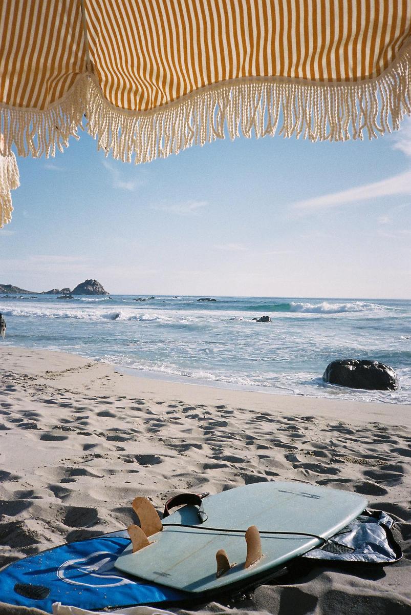 A surfboard rests on a sandy beach with an umbrella overhead and ocean waves in the background.