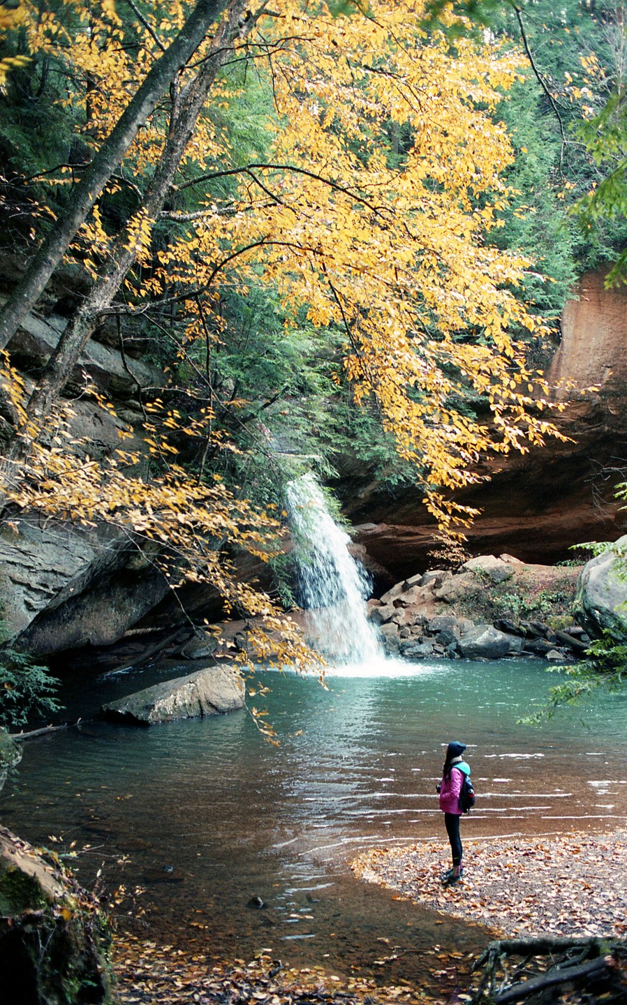 A person stands by a pool of water with a waterfall and autumn trees in the background.