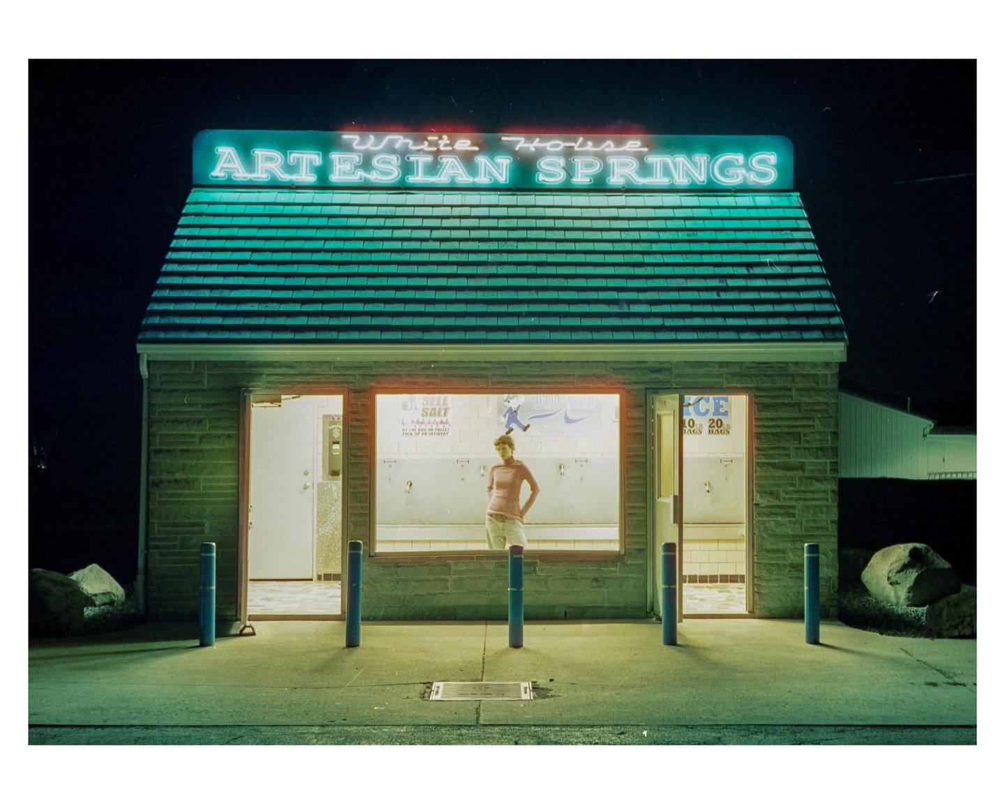 A woman stands inside a small building with a neon sign that reads "White House Artesian Springs" at night.